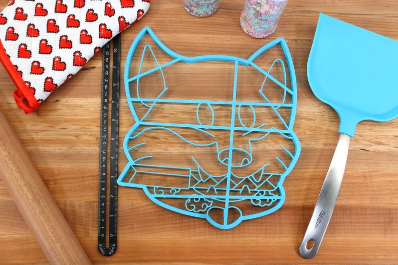 May include: A blue plastic cookie cutter shaped like a cartoon fox's face. The cutter is divided into sections for decorating cookies.