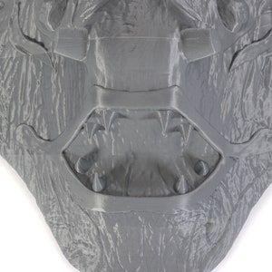 Puede incluir: Una escultura de pared gris texturizada, impresa en 3D, que representa la cara de un monstruo con la boca abierta y dientes afilados.