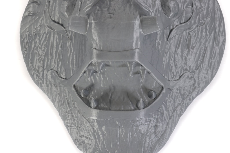 Puede incluir: Una escultura de pared gris texturizada, impresa en 3D, que representa la cara de un monstruo con la boca abierta y dientes afilados.