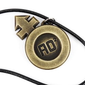 May include: A black cord necklace with a gold-toned pendant featuring a stylized arrow and a circular design with the text "AD2".