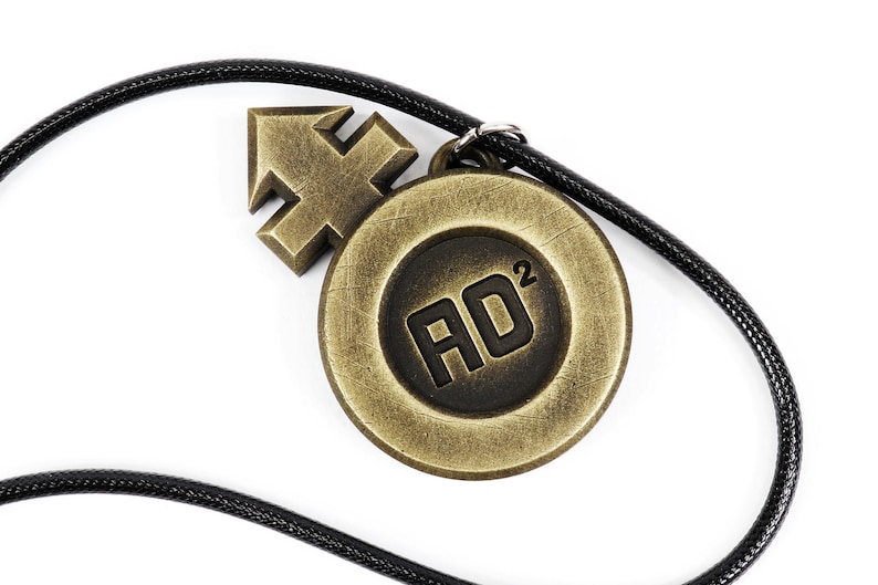 May include: A black cord necklace with a gold-toned pendant featuring a stylized arrow and a circular design with the text "AD2".