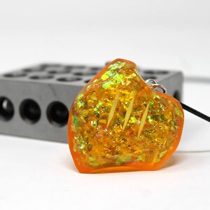 May include: A heart-shaped pendant necklace with a clear orange resin filled with gold and green glitter. The pendant is hanging from a black cord.