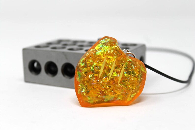 May include: A heart-shaped pendant necklace with a clear orange resin filled with gold and green glitter. The pendant is hanging from a black cord.