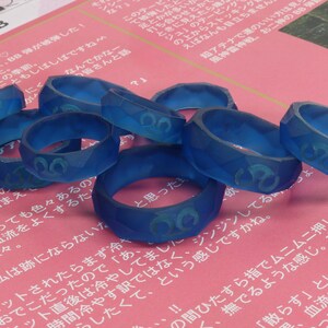 May include: A group of eight blue resin rings with a raised design. The rings are stacked on top of each other and are on a pink surface.