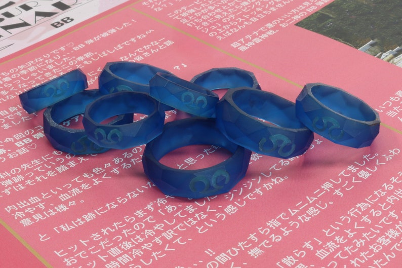 May include: A group of eight blue resin rings with a raised design. The rings are stacked on top of each other and are on a pink surface.