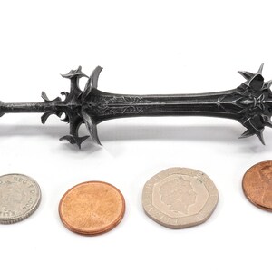 May include: A black metal sword with intricate details, shown next to three coins for scale.