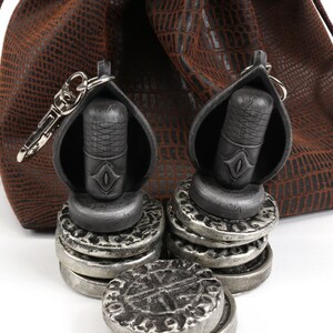 May include: A brown leather pouch with a drawstring closure and a silver metal key chain with a black metal charm. The charm is a small, cylindrical object with a raised design. The pouch is sitting on a white surface with a stack of silver coins.