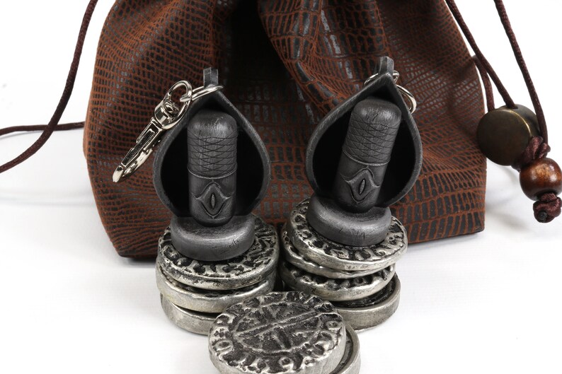 May include: A brown leather pouch with a drawstring closure and a silver metal key chain with a black metal charm. The charm is a small, cylindrical object with a raised design. The pouch is sitting on a white surface with a stack of silver coins.