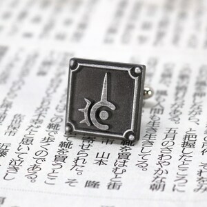 May include: Silver cufflinks with a square design featuring a stylized symbol with a circle and a spike. The symbol is on a dark gray background.