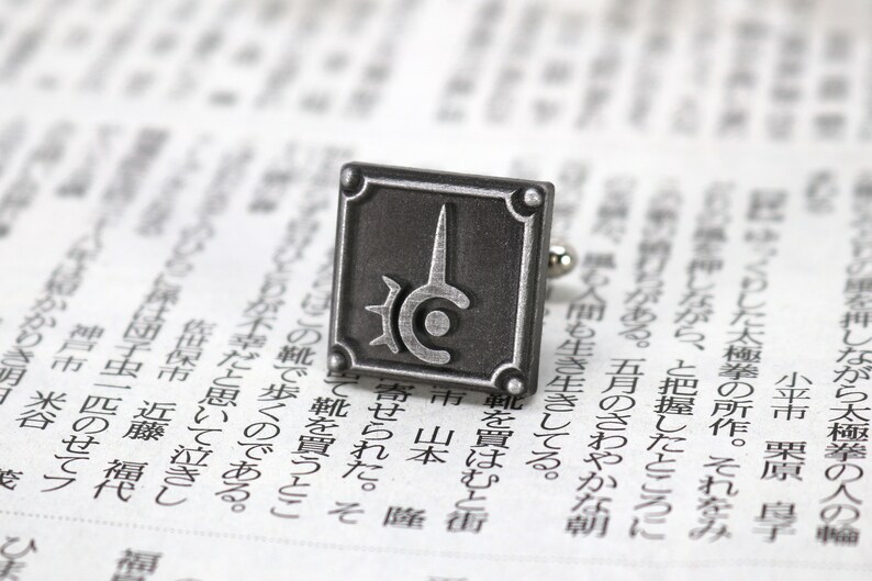 May include: Silver cufflinks with a square design featuring a stylized symbol with a circle and a spike. The symbol is on a dark gray background.