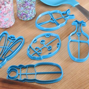 May include: Five blue plastic cookie cutters shaped like ladybugs, a beetle, and a butterfly on a wooden surface.