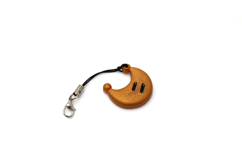 May include: A gold-colored crescent moon charm with black accents, attached to a black string with a silver clasp.