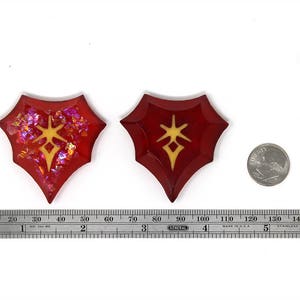 May include: Two red resin shield-shaped pendants with a gold star design. The pendants are approximately 5 cm in length. A US quarter is shown for scale.
