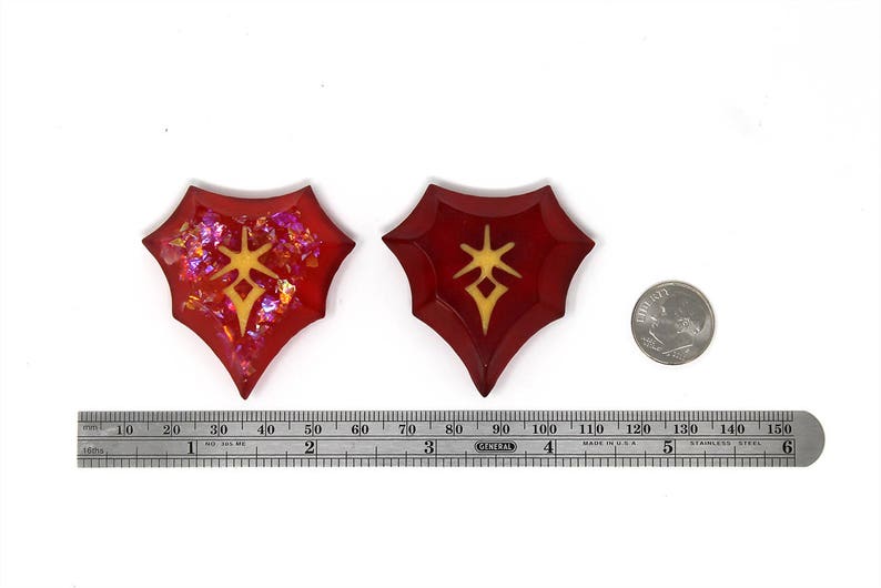 May include: Two red resin shield-shaped pendants with a gold star design. The pendants are approximately 5 cm in length. A US quarter is shown for scale.