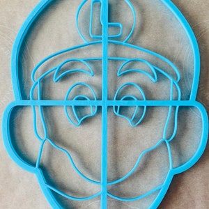 May include: A light blue plastic cookie cutter in the shape of a cartoon character's face. The cutter features the character's eyes, nose, mouth, and a circular emblem with the letter "L" on top of the head.