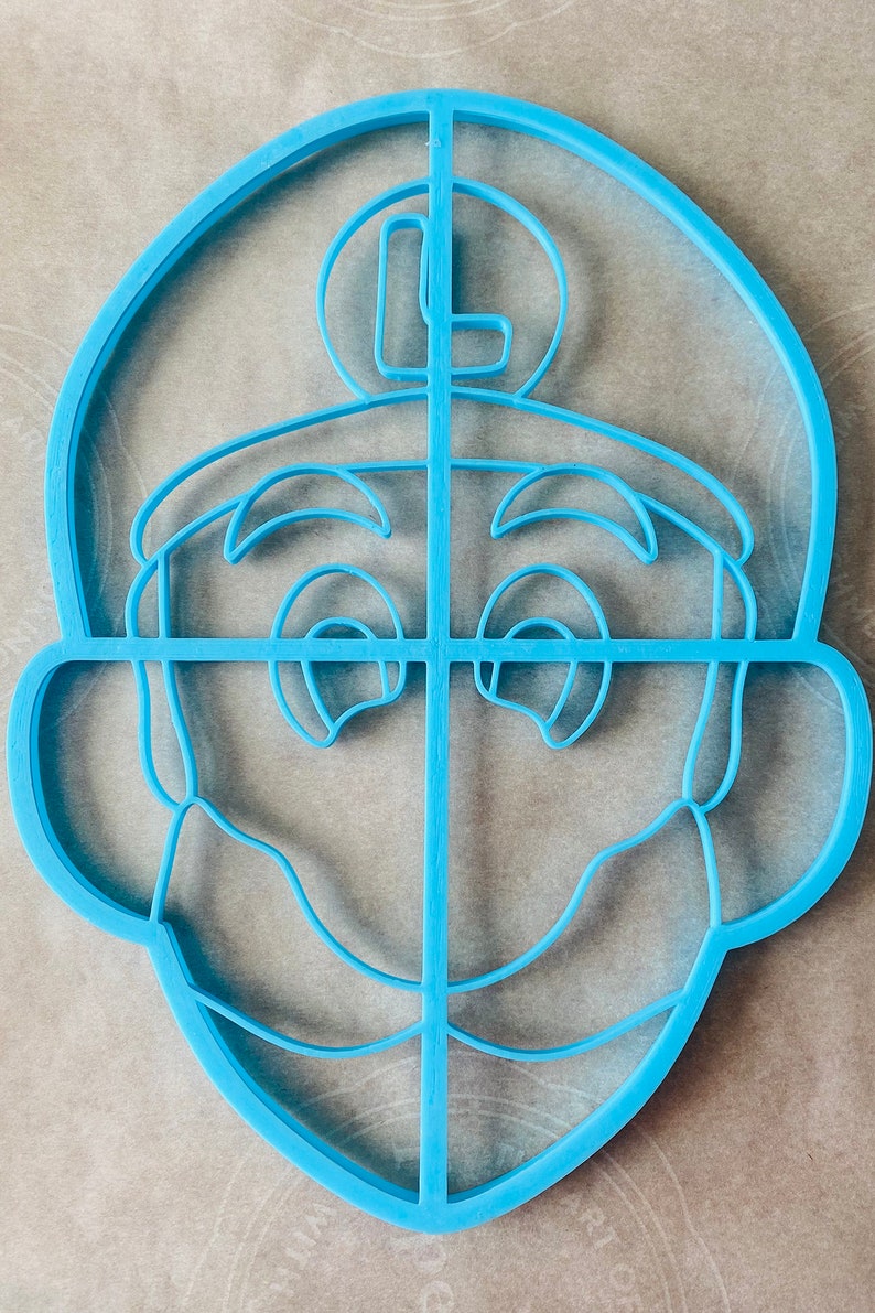 May include: A light blue plastic cookie cutter in the shape of a cartoon character's face. The cutter features the character's eyes, nose, mouth, and a circular emblem with the letter "L" on top of the head.