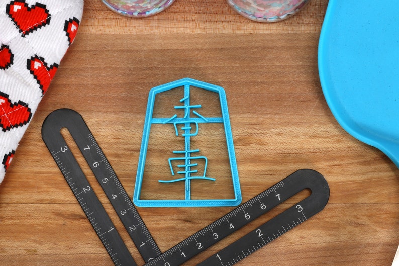 May include: A light blue plastic cookie cutter in the shape of a Japanese character, with black measuring tools and a blue spatula on a wooden surface. A white oven mitt with red pixelated hearts is in the background.