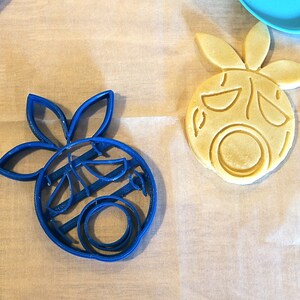 May include: Two cookie cutters in the shape of a cartoon face with a flower on top. One cutter is blue plastic and the other is made of yellow dough.