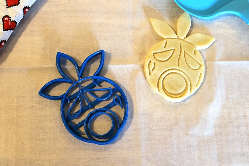 May include: Two cookie cutters in the shape of a cartoon face with a flower on top. One cutter is blue plastic and the other is made of yellow dough.