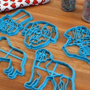 May include: A collection of light blue dog-shaped cookie cutters in various poses, including a dog's head, a dog wearing a hat, and a dog in a sitting position. The cutters are arranged on a wooden surface.