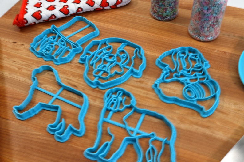 May include: A collection of light blue dog-shaped cookie cutters in various poses, including a dog's head, a dog wearing a hat, and a dog in a sitting position. The cutters are arranged on a wooden surface.