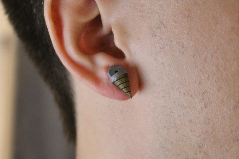 May include: A single stud earring shaped like a drill bit, with a gray top and a bronze-colored, cone-shaped drill. The earring is worn in an ear piercing.