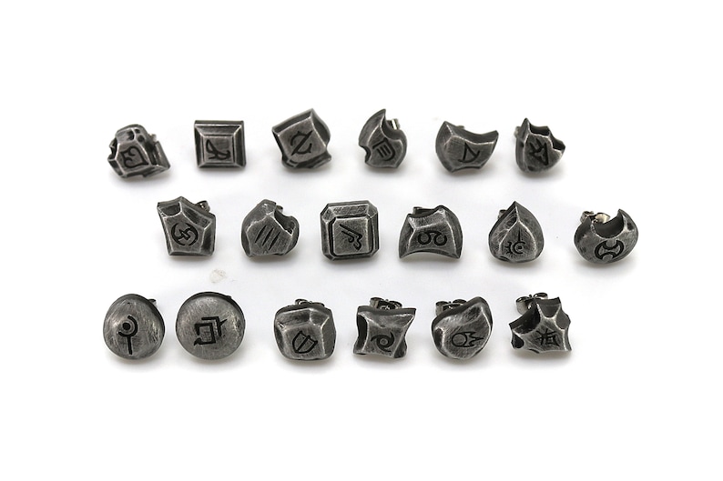 May include: A set of 18 silver-toned metal earrings with various designs. Each earring features a different symbol or design, including geometric shapes, abstract patterns, and stylized characters.