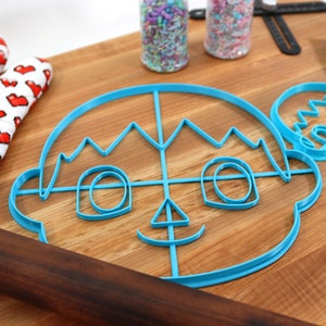 May include: Two blue plastic cookie cutters shaped like a cartoon face with a crown and a smaller version of the same face. The cutters are on a wooden cutting board with a rolling pin and a red and white oven mitt.