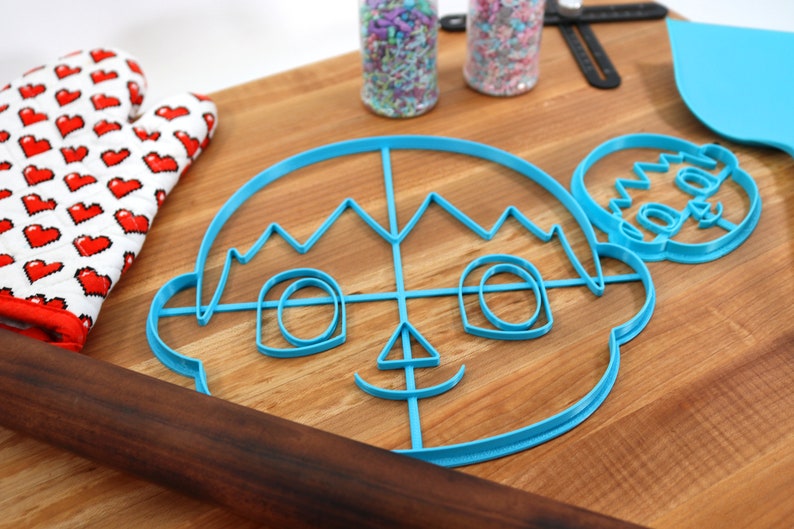 May include: Two blue plastic cookie cutters shaped like a cartoon face with a crown and a smaller version of the same face. The cutters are on a wooden cutting board with a rolling pin and a red and white oven mitt.