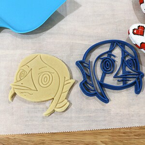May include: Two cookie cutters, one blue and one beige, shaped like a cartoon character with large eyes and a wide mouth. The cutters are on a white surface.
