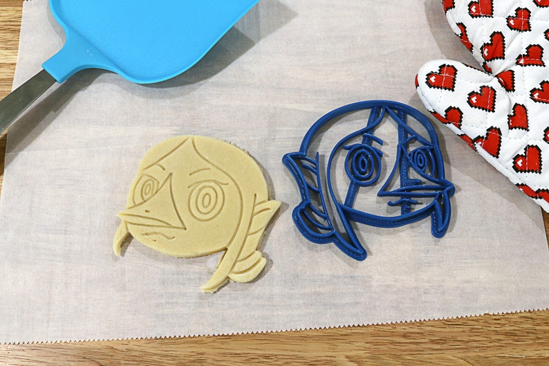 May include: Two cookie cutters, one blue and one beige, shaped like a cartoon character with large eyes and a wide mouth. The cutters are on a white surface.