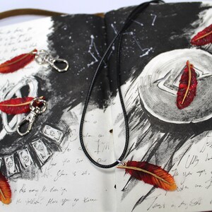 May include: A black leather cord necklace with a red and orange feather pendant. The necklace is laying on a notebook with a black and white drawing of a moon and stars. The notebook also has a few red and orange feather keychains.