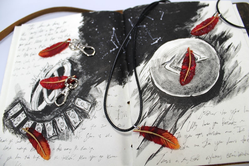May include: A black leather cord necklace with a red and orange feather pendant. The necklace is laying on a notebook with a black and white drawing of a moon and stars. The notebook also has a few red and orange feather keychains.