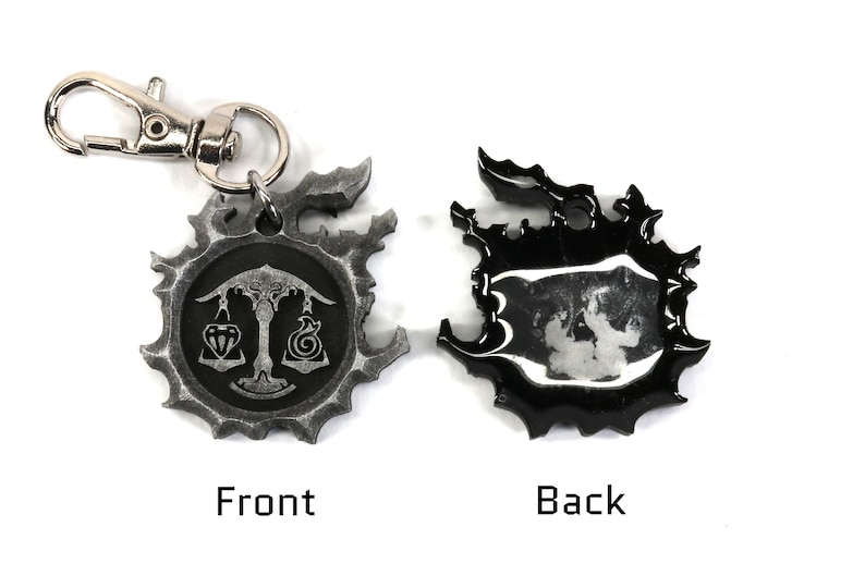 May include: A silver metal keychain with a black and white design. The front features a scale with a diamond and a spiral design. The back is black with a silver design.