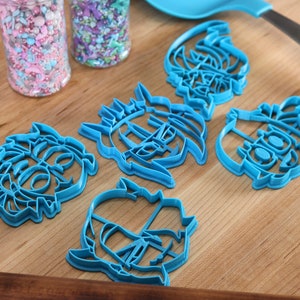 May include: Five blue plastic cookie cutters shaped like cartoon faces. The cutters are on a wooden surface.