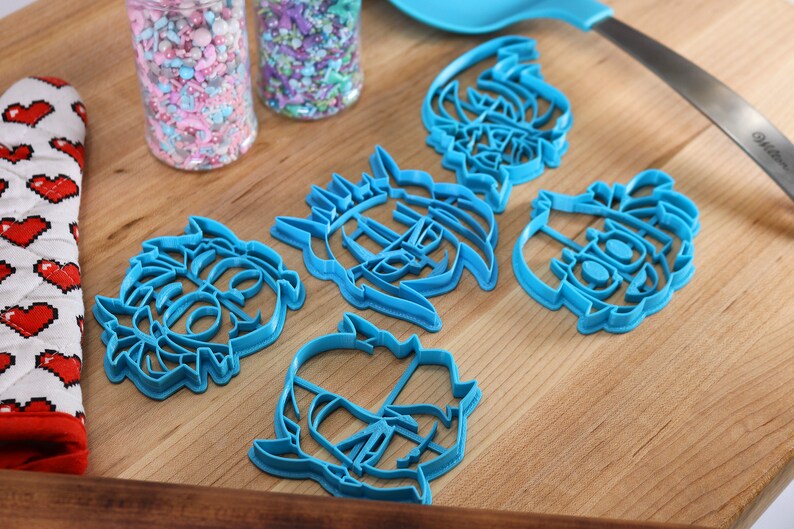May include: Five blue plastic cookie cutters shaped like cartoon faces. The cutters are on a wooden surface.