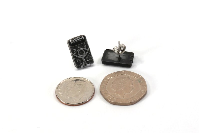 May include: A pair of black and silver earrings featuring a stylized eye design. The earrings are shown next to a US dime for size comparison.