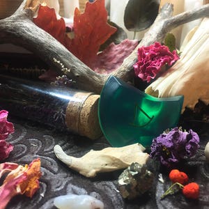 May include: A green glass rune with a gold symbol on a dark background with dried flowers, a bone, and a glass bottle with a cork stopper.