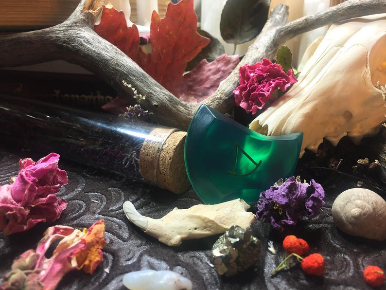May include: A green glass rune with a gold symbol on a dark background with dried flowers, a bone, and a glass bottle with a cork stopper.