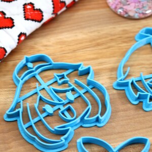 May include: Two blue plastic cookie cutters shaped like cartoon characters. The cutters are on a wooden surface.