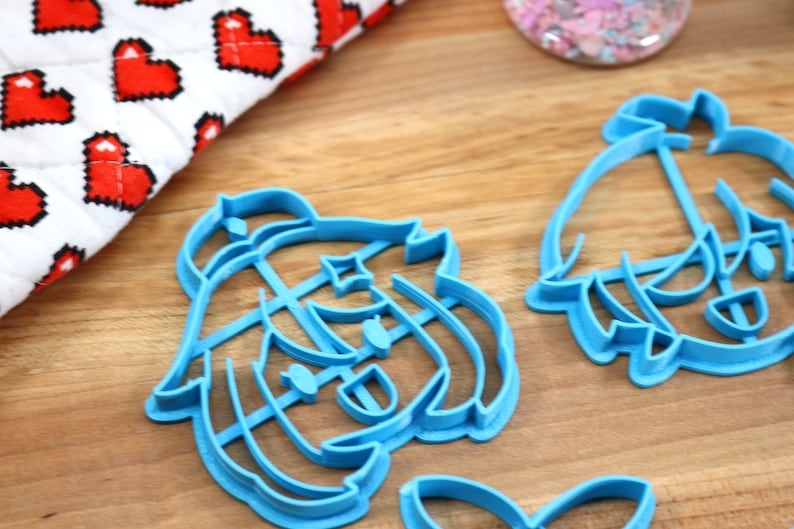 May include: Two blue plastic cookie cutters shaped like cartoon characters. The cutters are on a wooden surface.