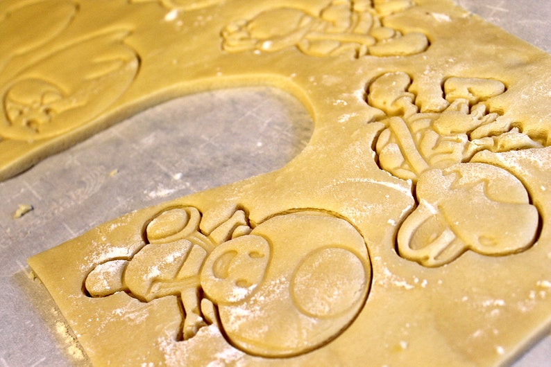 May include: Close-up of a cookie sheet with raw sugar cookie dough that has been cut out into various shapes. The shapes include a mushroom, a star, and a character from a video game.