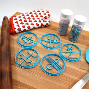 May include: A wooden rolling pin, a blue silicone spatula, a pair of red heart-patterned oven mitts, and five blue cookie cutters with geometric designs are arranged on a wooden cutting board. Two jars of colorful sprinkles are also on the board.