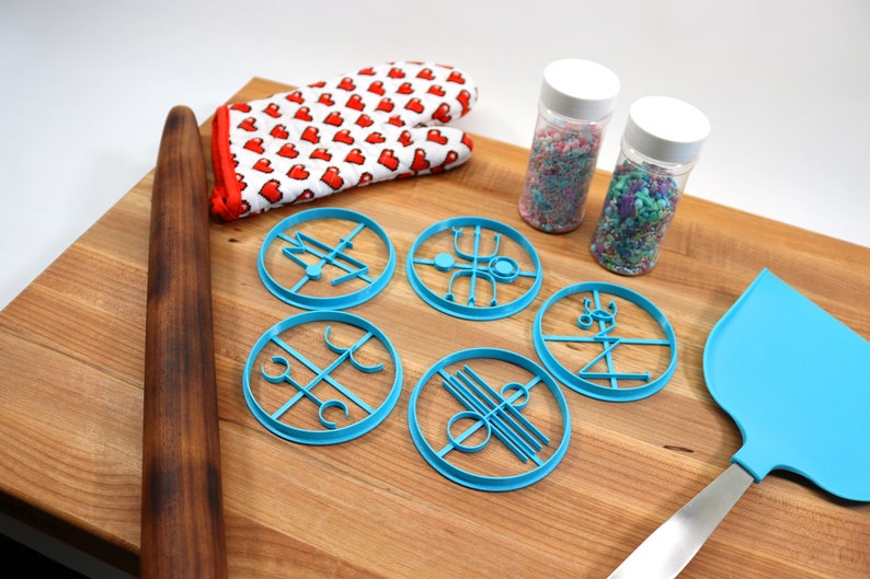 May include: A wooden rolling pin, a blue silicone spatula, a pair of red heart-patterned oven mitts, and five blue cookie cutters with geometric designs are arranged on a wooden cutting board. Two jars of colorful sprinkles are also on the board.