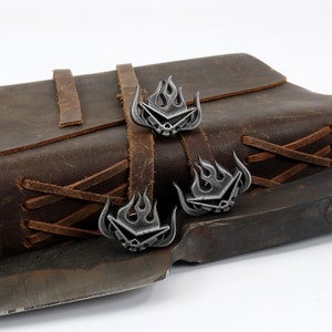 May include: Three silver-toned metal bookmarks with a flame design and a skull in the center. The bookmarks are attached to a brown leather journal with brown leather straps.