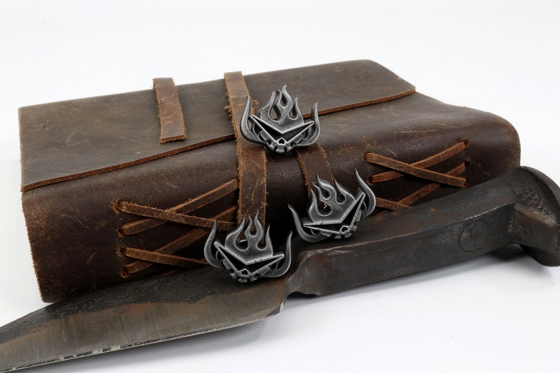 May include: Three silver-toned metal bookmarks with a flame design and a skull in the center. The bookmarks are attached to a brown leather journal with brown leather straps.