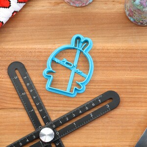 May include: A blue plastic cookie cutter shaped like a cartoon character with a surprised expression. The cutter is on a wooden surface.