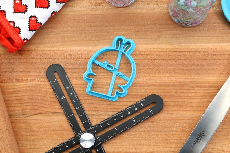May include: A blue plastic cookie cutter shaped like a cartoon character with a surprised expression. The cutter is on a wooden surface.