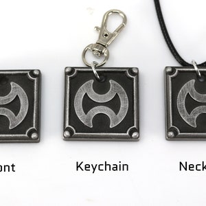 May include: A silver-colored metal pendant with a black and silver design. The pendant is available as a keychain or necklace.