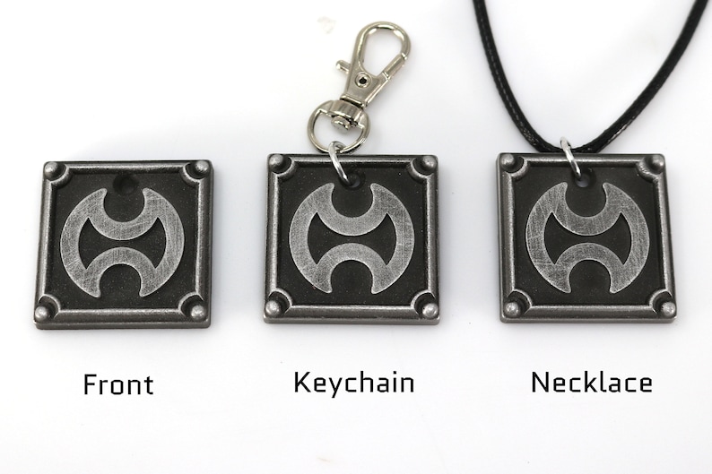 May include: A silver-colored metal pendant with a black and silver design. The pendant is available as a keychain or necklace.